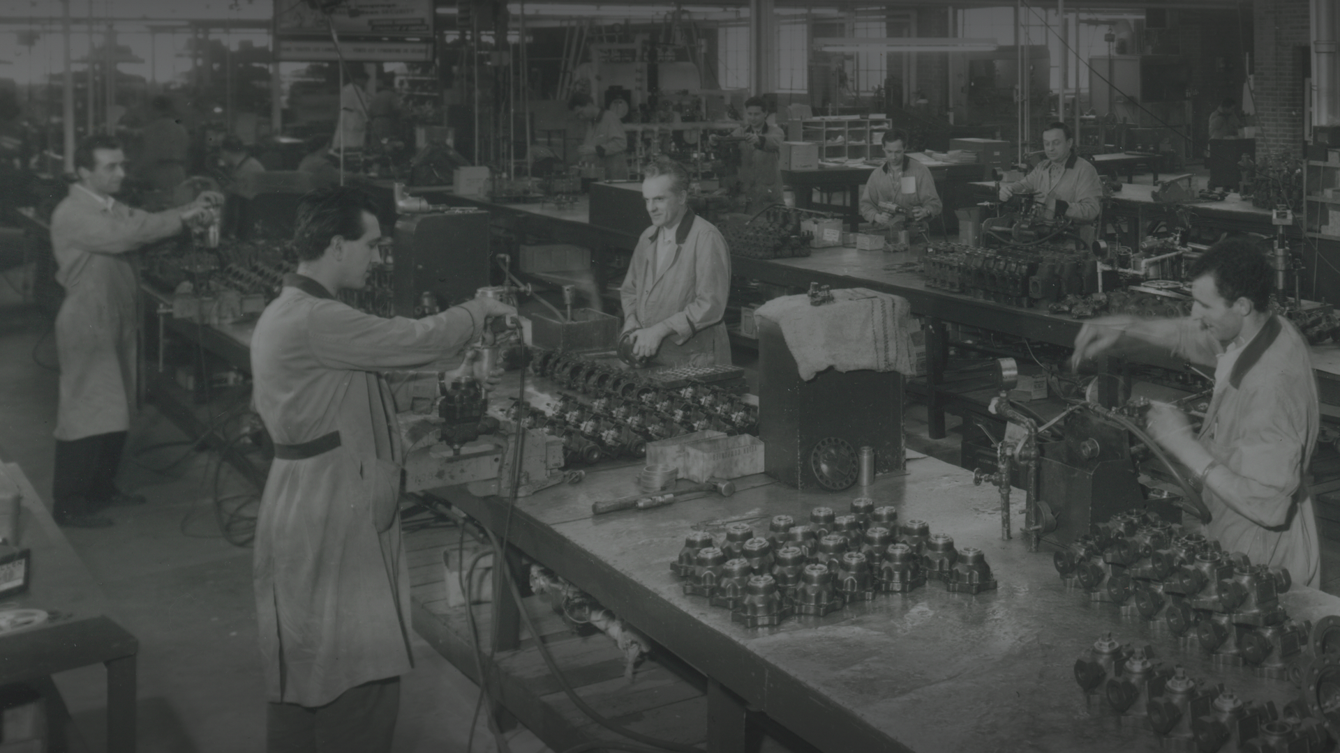 A black-and-white photo of a busy factory floor during the mid-20th century. Workers in lab coats are seen assembling and working on various mechanical parts at long, cluttered workbenches lined with tools and components under a well-lit industrial environment.