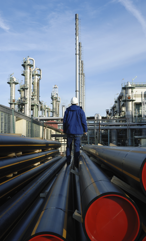 A person in a blue work uniform and white hard hat stands on large industrial pipes, looking towards an oil refinery complex with tall stacks and interconnected machinery in the background under a clear sky.