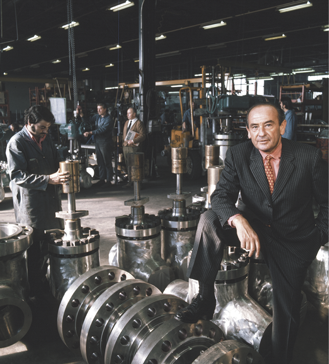 A man in a suit with a pink tie stands with one foot raised on large metal equipment inside a factory; several workers can be seen in the background working on machinery and equipment amidst industrial surroundings.
