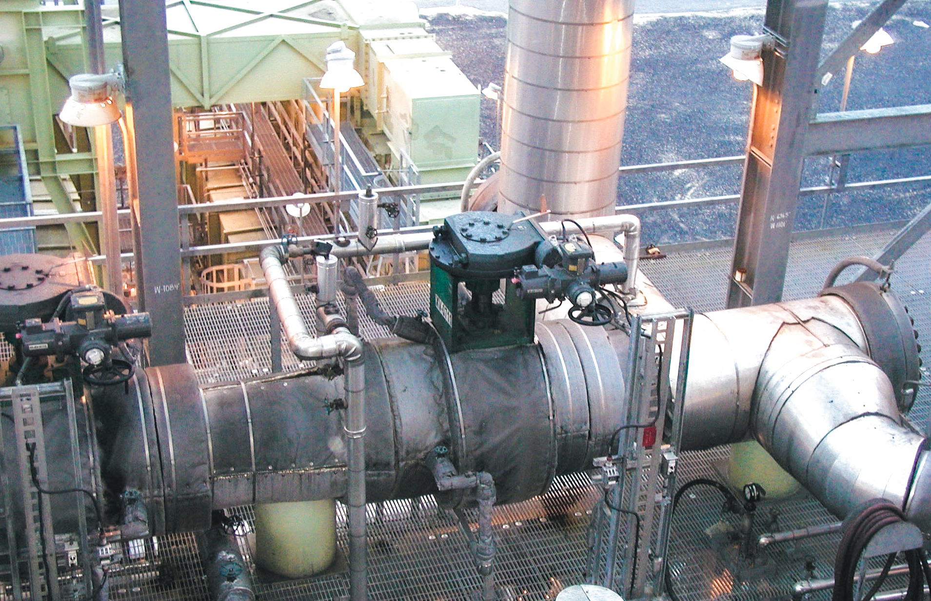 Large industrial pipes and valves in a factory or processing plant, with metal walkways, railings, and overhead lights visible. The area is clean and organized, with various equipment and machinery in the background.