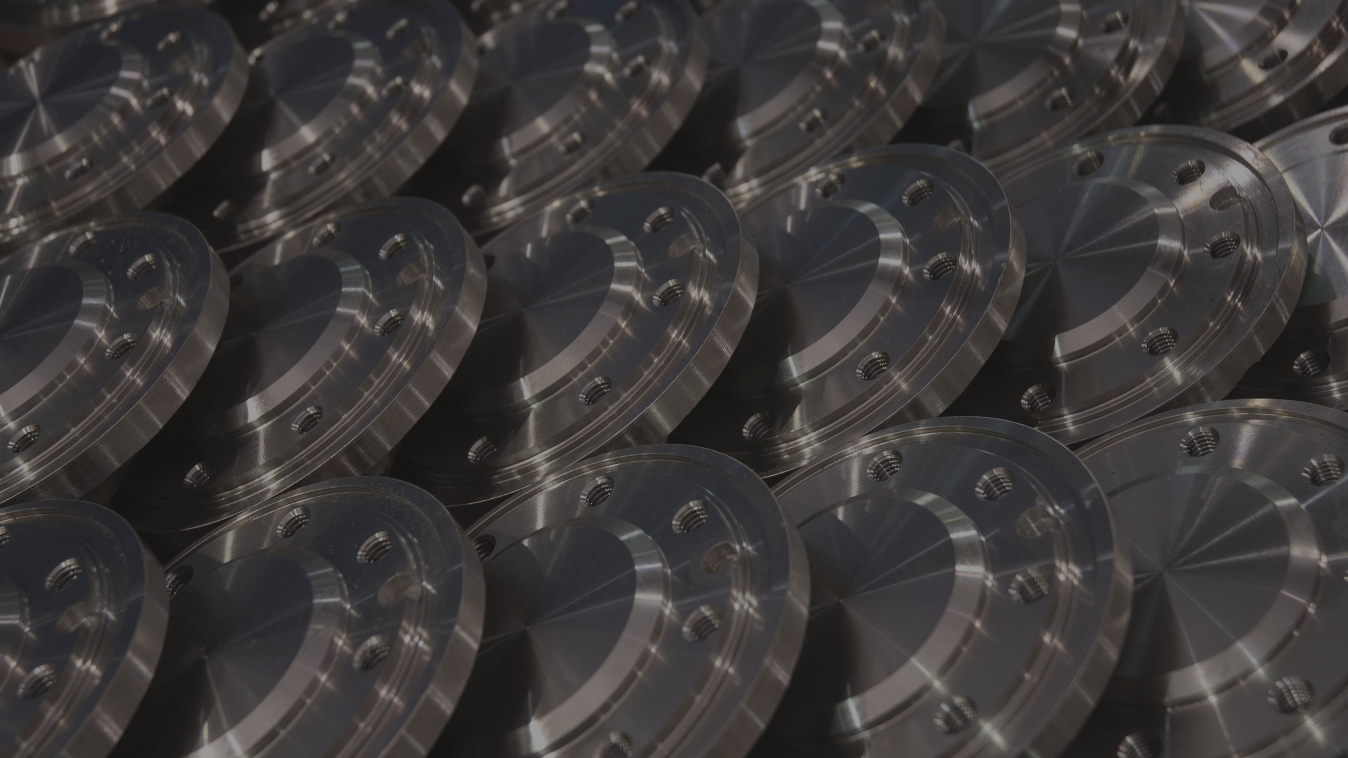 A close-up of multiple shiny, metallic circular machine parts arranged in a grid pattern. The parts have several holes and reflective, smooth surfaces, displaying intricate machining details and uniformity. The lighting highlights their polished texture.