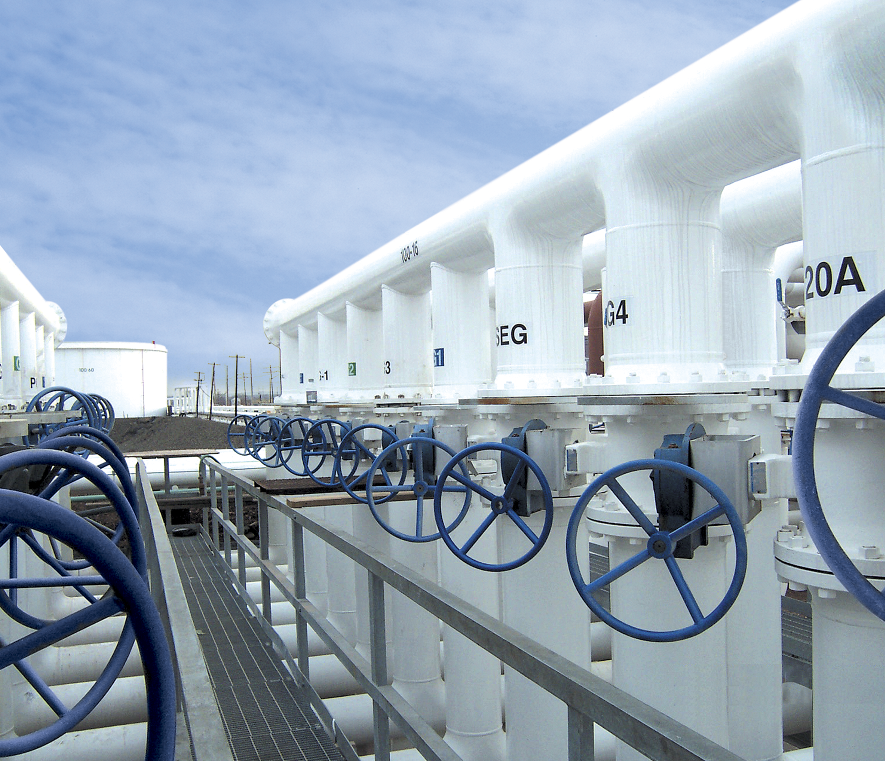 A series of large white pipes with blue wheel valves are aligned in an industrial facility with a clear blue sky in the background. The pipes are labeled with numbers and letters, and metal railings run alongside the system on a grated walkway.