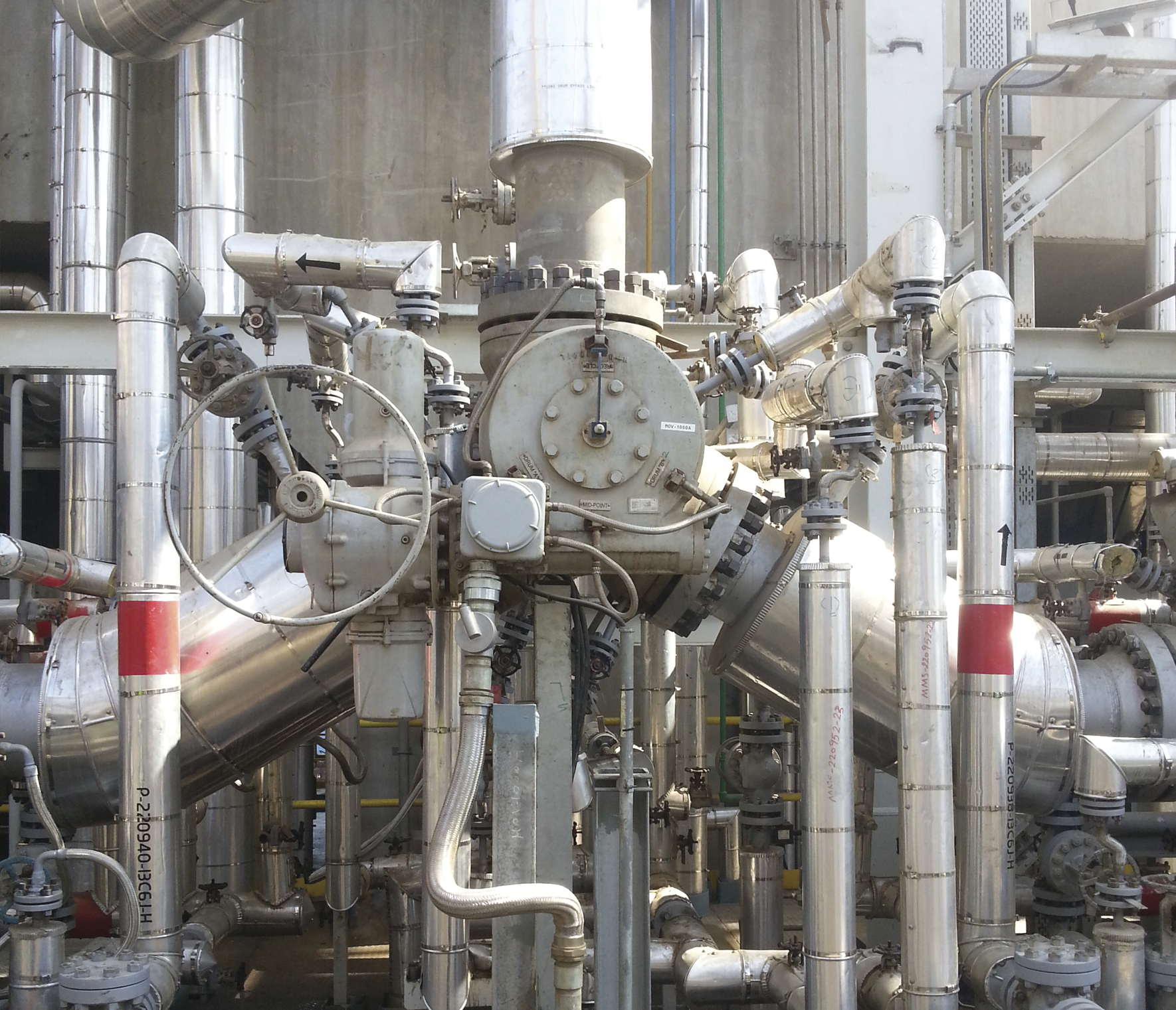 A complex assembly of industrial pipes and valves is visible, featuring a large central valve surrounded by numerous interconnected metallic pipes and control systems. The setup is part of a large-scale mechanical or chemical processing plant.