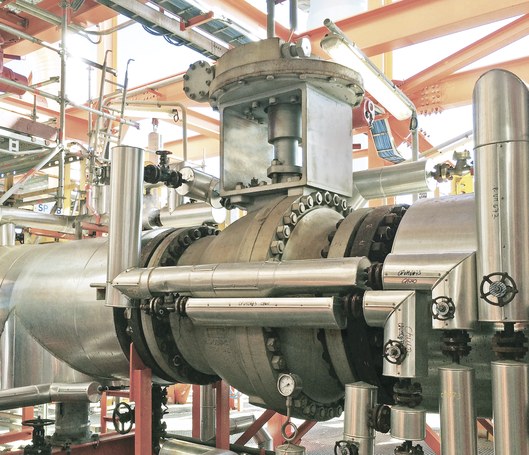A close-up view of industrial piping and equipment in a manufacturing or processing facility. The setup includes large steel pipes, valves, flanges, and various metal fittings, with an orange structural framework in the background.